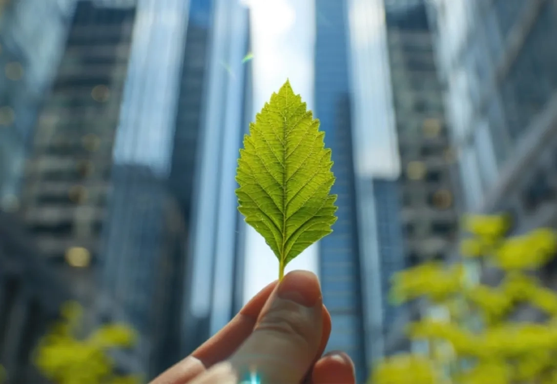 A hand holding a fresh green leaf against a backdrop of commercial office spaces, highlighting Good Earth Infra's commitment to sustainability in its LEED Gold certified developments and sustainable leasing practices.